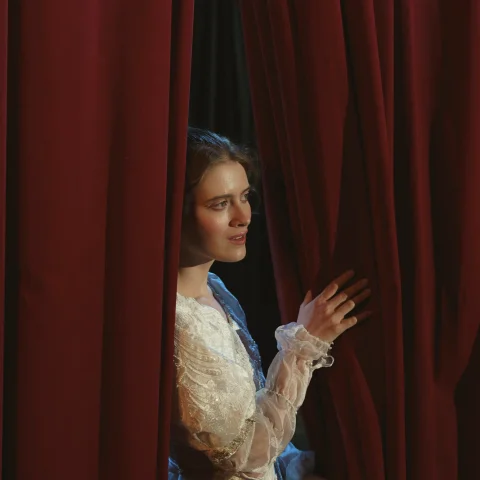 A woman on a theatre stage having a look from the red curtain
